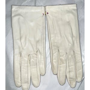 May include: A pair of ivory-colored leather gloves. The gloves have a classic design with a smooth texture and visible stitching. The gloves are laid flat, showcasing their shape and details. The gloves are a neutral color, suitable for various outfits.
