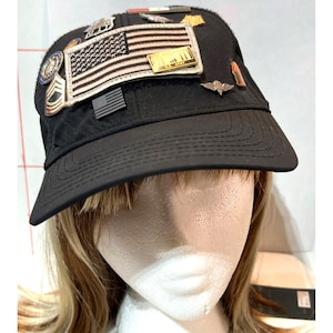 May include: A black baseball cap adorned with various military-style pins and patches, including a prominent embroidered American flag patch, a small black flag emblem, and several metallic insignia pins attached to the front and crown.