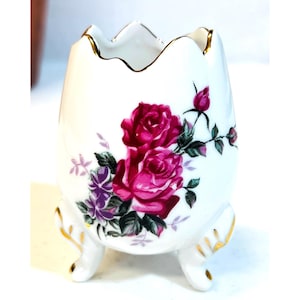 May include: White ceramic egg-shaped vase with a floral design featuring pink roses and purple accents. The vase has three small feet and gold trim along the scalloped rim and feet. A decorative piece for home decor.
