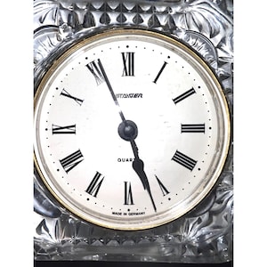 May include: A clear crystal quartz clock with a white face and black Roman numerals. The clock face has a gold-coloured rim and the hands are black. The clock is marked "STAGER" and "QUARTZ" and "MADE IN GERMANY".