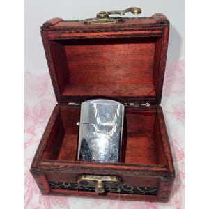 May include: A silver-toned metal lighter with a visible engraved logo sits inside an open, rectangular wooden box. The box features a dark reddish-brown finish, a hinged lid, and a brass-colored metal latch mechanism on the front exterior.