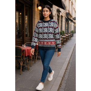 Puede incluir: Una mujer con un suéter azul marino con un patrón geométrico en rojo y blanco, combinado con jeans azules y zapatillas blancas. El suéter tiene cuello redondo y mangas largas. El fondo muestra una escena de calle europea.