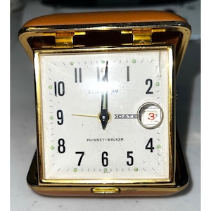 May include: A vintage Phinney-Walker travel alarm clock. The clock has a square face with black numerals and gold hands. The date is visible in a small window. The clock is encased in a brown leather-like case with gold trim.