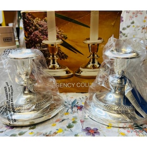May include: Two silver-colored candle holders with a detailed base and a cup-shaped top. The candle holders are in plastic bags. The background includes a box with a floral design and the text "REGENCY COLL".