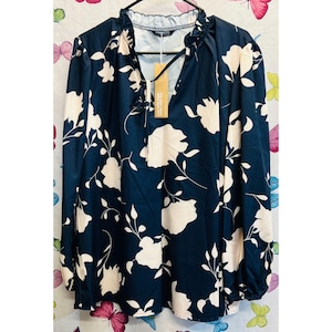 May include: Navy blue long-sleeved blouse with a cream floral pattern. The blouse has a V-neck with a ruffled collar and tie detail. A tag is attached to the neckline.