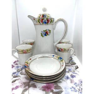 Puede incluir: Un juego de café de porcelana blanca que incluye una cafetera alta, cuatro tazas a juego y una pila de cuatro platillos. Cada pieza está decorada con motivos florales pintados a mano en azul, rosa y verde, con un fino borde dorado.