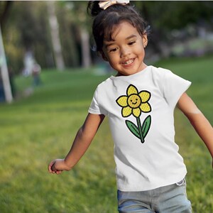 May include: A young person wearing a white t-shirt with a yellow flower graphic. The flower has a smiling face and green leaves. The person is standing in a grassy field.