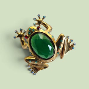 May include: A gold-toned frog brooch featuring a large, oval, faceted dark green central stone. The frog's body is accented with small, sparkling blue and red rhinestones embedded along its limbs, head, and back, creating a detailed, textured appearance.