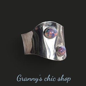 Peut inclure: Un bracelet manchette en argent avec deux pierres d'opale irisées serties dans le métal. Le bracelet est incurvé et présente un design ondulé.