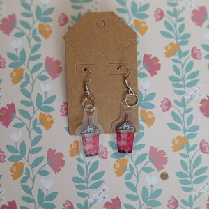 May include: A pair of dangle earrings featuring a clear acrylic design of a pink drink with whipped cream. The earrings hang from silver-colored hooks and are displayed on a brown tag against a floral background.