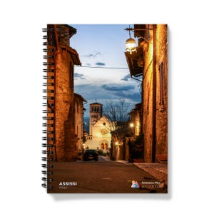 May include: Spiral-bound notebook featuring a photograph of a street in Assisi, Italy, at dusk. The image shows stone buildings, streetlights, and a church in the distance. The text "ASSISI ITALY" is printed on the bottom left.