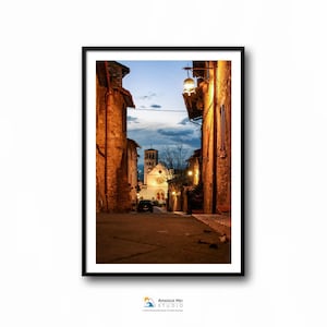 May include: A narrow, cobblestone street in a European town at dusk. The street is lined with old, stone buildings, and a church is visible in the distance. The street is lit by a single street lamp.