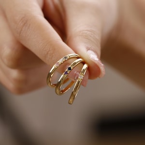 May include: Three gold rings with colourful gemstones. Each ring has a row of small gemstones in different colours, including pink, purple, and white.