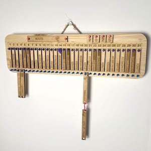 Op de afbeelding: Een houten kalender met een schuifsysteem om dagen te markeren. De kalender is voor de maand maart 2025. De dagen van de week zijn in het Frans gelabeld: Lundi, Mardi, Mercredi, Jeudi, Vendredi, Samedi, Dimanche.