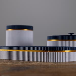 Puede incluir: Tres contenedores cilíndricos blancos con tapa azul y una banda dorada en el medio. Los contenedores están sobre una superficie de madera.