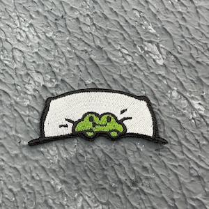May include: A white and black embroidered patch featuring a green frog peeking out from behind a white banner. The frog has a black outline and a simple smiling face.