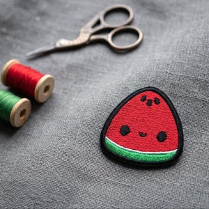 May include: A triangular watermelon patch featuring a red centre, a green rind, and a black border. The patch has embroidered black eyes and a small mouth, creating a face. It is placed on grey fabric near red and green thread spools and scissors.