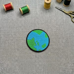 May include: A round embroidered patch depicting the Earth with blue oceans and green continents, outlined in black. Several spools of thread in red, green, yellow, and black are scattered around the patch, along with a pair of gold scissors. The patch is on a gray fabric.