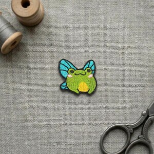 May include: An embroidered patch featuring a green frog with blue wings, a yellow belly, and small pink cheeks. The patch is positioned between a wooden spool of thread and a pair of metal scissors on a textured grey fabric surface.