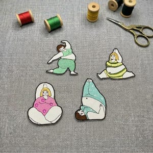 May include: Four embroidered patches depicting figures in yoga poses, in green, pink, and blue. The patches are on a gray surface with spools of thread and gold scissors. The patches are in the shape of the figures.