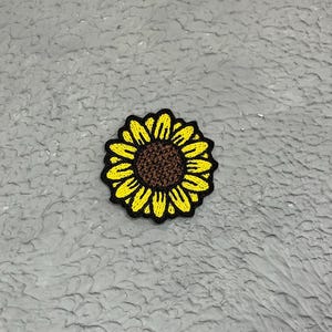 May include: A yellow embroidered sunflower patch with a brown centre. The patch is on a grey textured background.