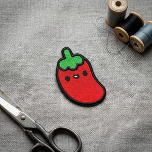 Peut inclure: Écusson brodé en forme de piment rouge avec une tige verte et un contour noir. Le patch présente un visage mignon avec des yeux en points noirs et une petite bouche blanche. Sur un tissu de lin gris, avec des bobines de fil et des ciseaux en arrière-plan.