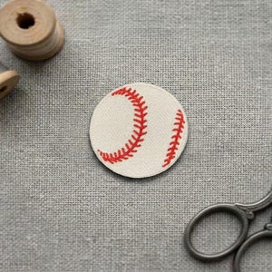 May include: A baseball patch with red stitching on a white background. The patch is round and features the classic baseball design. A pair of scissors and two spools of thread are also in the image.