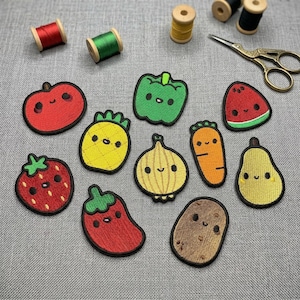 Op de afbeelding: Een verzameling geborduurde patches met verschillende lachende groenten en fruit. De patches bevatten een rode appel, groene paprika, gele ananas, oranje wortel en een bruine aardappel. Ook zijn er klosjes garen en een gouden schaar aanwezig.