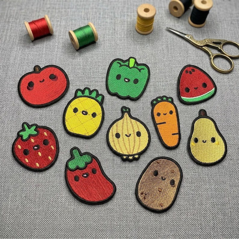 Op de afbeelding: Een verzameling geborduurde patches met verschillende lachende groenten en fruit. De patches bevatten een rode appel, groene paprika, gele ananas, oranje wortel en een bruine aardappel. Ook zijn er klosjes garen en een gouden schaar aanwezig.