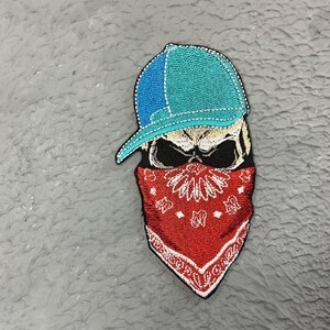 May include: Embroidered patch of a skull wearing a blue baseball cap and a red bandana. The skull has black eyes and a white face.