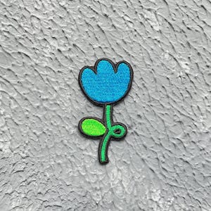 Peut inclure: Un patch brodé en forme de fleur bleu et vert. La fleur est de style cartoon simple avec une forme arrondie et une seule feuille.