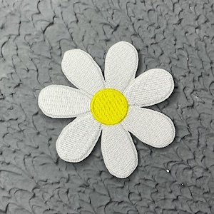 Peut inclure: Une fleur de marguerite en feutre blanc avec un centre jaune. La fleur est sur un fond texturé gris.