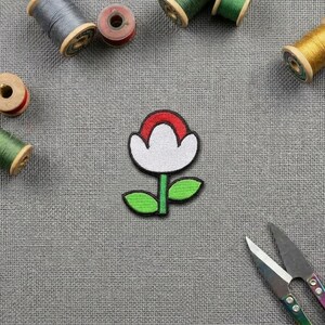 May include: A fabric patch shaped like a stylised flower with a white centre, a red top arch, and a green stem with two leaves. The patch is surrounded by various wooden and plastic spools of sewing thread and a pair of metal sewing scissors.