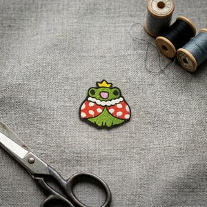 May include: An embroidered patch featuring a green frog wearing a yellow crown and a red cape with white polka dots. The frog has black eyes and a pink mouth. The patch is positioned on a grey fabric surface near metal scissors and thread spools.