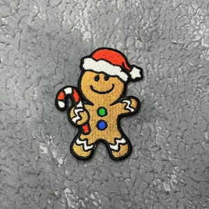 Peut inclure: Un bonhomme en pain d'épices portant un chapeau de Père Noël rouge et blanc tient une canne en sucre d'orge dans sa main droite. Il sourit et a des boutons verts et bleus sur son corps en pain d'épices.
