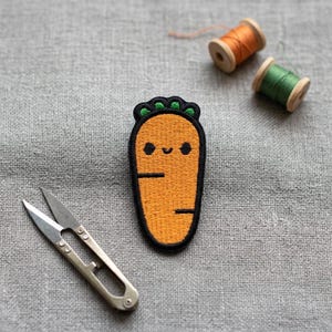 May include: An embroidered orange carrot patch with a smiling face and black outline. The patch is next to small scissors and two spools of thread, one orange and one green, on a gray fabric background.