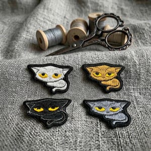 May include: Four embroidered cat patches in white, gold, black, and grey, each featuring bright yellow eyes. Above the patches lie a pair of ornate, dark metal embroidery scissors and three wooden spools wrapped in grey thread on a textured fabric.
