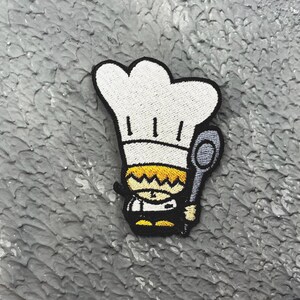 May include: Embroidered patch featuring a cartoon chef wearing a white hat and holding a spoon. The chef has yellow hair and a black outline. The patch is on a textured gray surface.