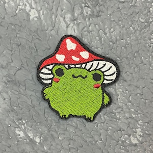 Peut inclure: Patch brodé représentant une grenouille verte portant un chapeau de champignon rouge et blanc. La grenouille a des yeux noirs et un sourire blanc.