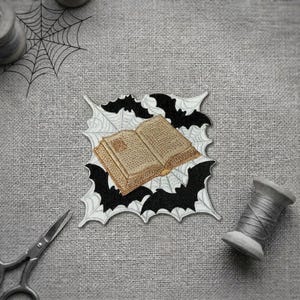 Peut inclure: Un écusson brodé représentant un livre ouvert de couleur beige avec des pages visibles, centré sur une toile d'araignée blanche et entouré de silhouettes noires de chauves-souris en vol. Le motif est carré avec des bords dentelés.