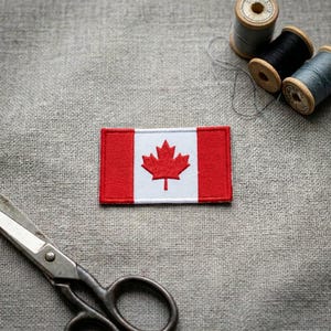 May include: A rectangular embroidered patch featuring the Canadian flag with a red maple leaf centered on a white background flanked by two red vertical bars. The patch is positioned near metal scissors and three spools of sewing thread.