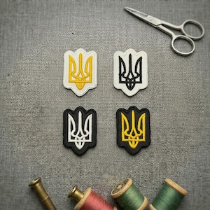 May include: Four embroidered patches featuring the Ukrainian Tryzub emblem. Two patches have white backgrounds with yellow and black stitching, while two have black backgrounds with white and yellow stitching. Metal scissors and thread spools are present.