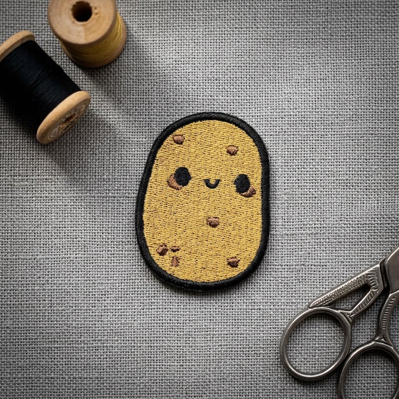 Op de afbeelding: Een geborduurde patch in de vorm van een lachende aardappel, geel met een zwarte rand. De aardappel heeft zwarte ogen en een gebogen mond, met bruine vlekken. Naaibenodigdheden, waaronder garenklosjes en een schaar, staan op de achtergrond.