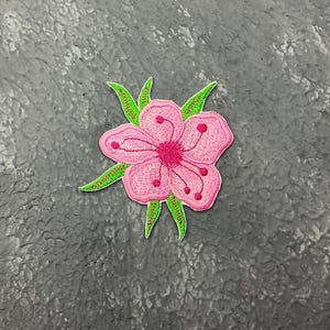 May include: Embroidered pink flower patch with green leaves. The flower has a dark pink centre and small red dots. The patch is likely for clothing or accessories.