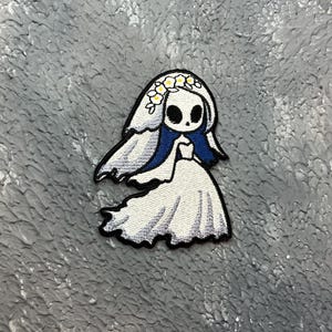 May include: Embroidered patch of a cartoon bride with a skull face, wearing a white dress and veil with blue accents. The bride has a floral crown. The patch is on a textured grey surface.