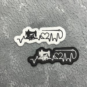 May include: Two embroidered patches, one white and one black, with a sewing machine, floral design, heart, and a heartbeat line. The patches are on a textured grey surface.