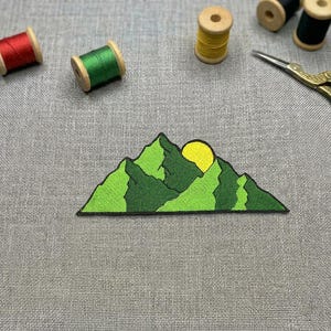 May include: A green and black embroidered mountain patch featuring a yellow sun. The image also shows spools of red, green, yellow, and black thread, along with gold scissors. The patch design is detailed.
