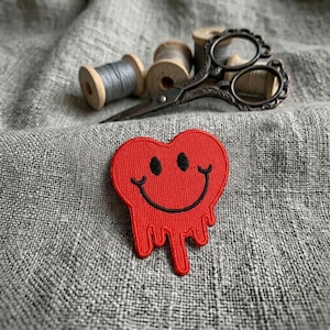 May include: A red heart-shaped patch featuring a black embroidered smiley face with a dripping effect. The patch is positioned near vintage-style metal scissors and several wooden spools of grey thread, all resting on a textured grey fabric surface.