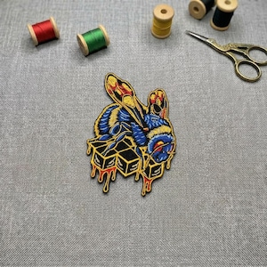 May include: An embroidered patch featuring a blue and yellow bee resting on a honeycomb structure with dripping honey. The patch is surrounded by two small spools of red and green thread, two wooden spools, and a pair of gold metal scissors.