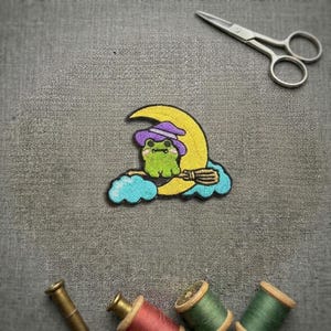 Peut inclure: Un écusson brodé représentant une grenouille verte portant un chapeau de sorcière violet, assise sur un croissant de lune jaune avec un balai marron, au-dessus de nuages bleu clair. L'écusson est présenté avec des ciseaux et des bobines de fil.